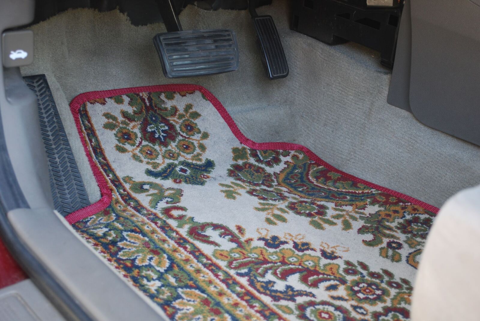 Close-up of the driver's mat showing the sewn red webbing binding, with the rug's border running diagonally